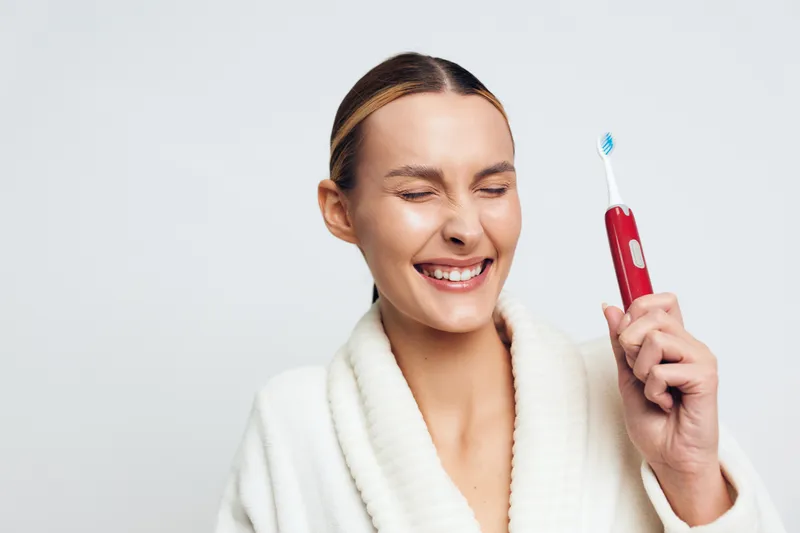 Smiling woman in white bathrobe holds electric toothbrush with closed eyes showing happiness and fresh morning routine isolated on white background with clean minimal style.