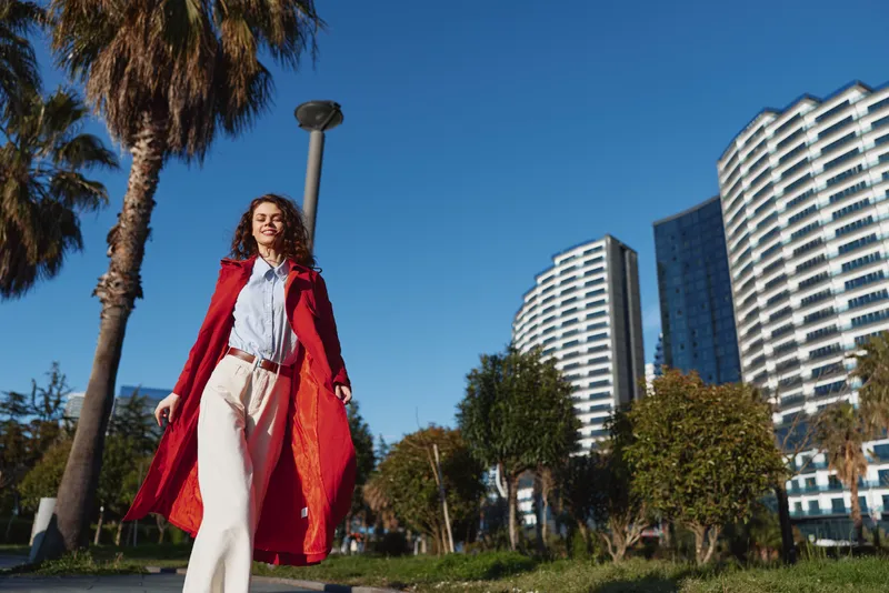 Young woman with curly hair walks confidently outdoors wearing a long red coat and white pants on a sunny day with modern buildings in the background.