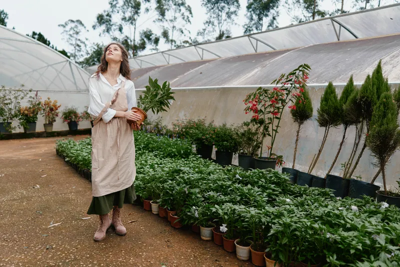 Young woman in a greenhouse smiling while holding a potted plant, dressed in casual attire with a light apron, surrounded by lush greenery and vibrant flowers