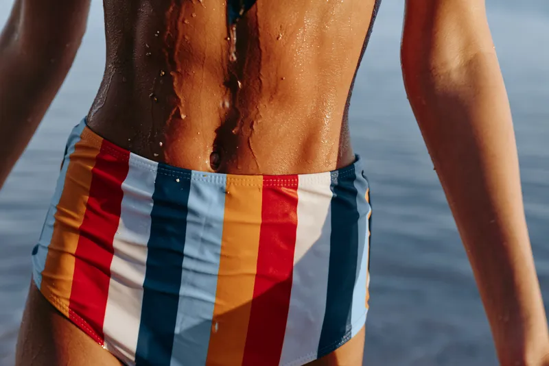 Close up view of wet skin and swimwear, striped trunks on a beach with ocean water in the background, sunlight reflection creates a summer vacation feeling, outdoor relaxation and fitness lifestyle