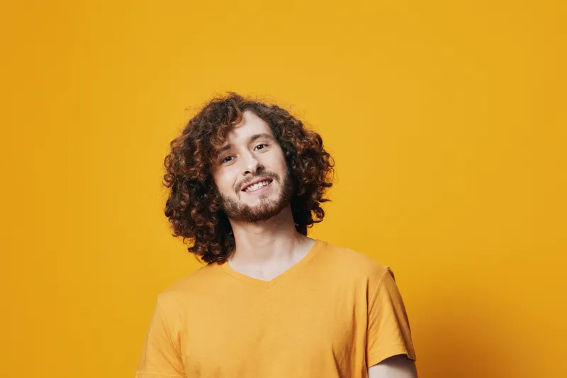 Smiling young man with curly hair in a yellow t shirt against a bright yellow background, conveying joy and positivity
