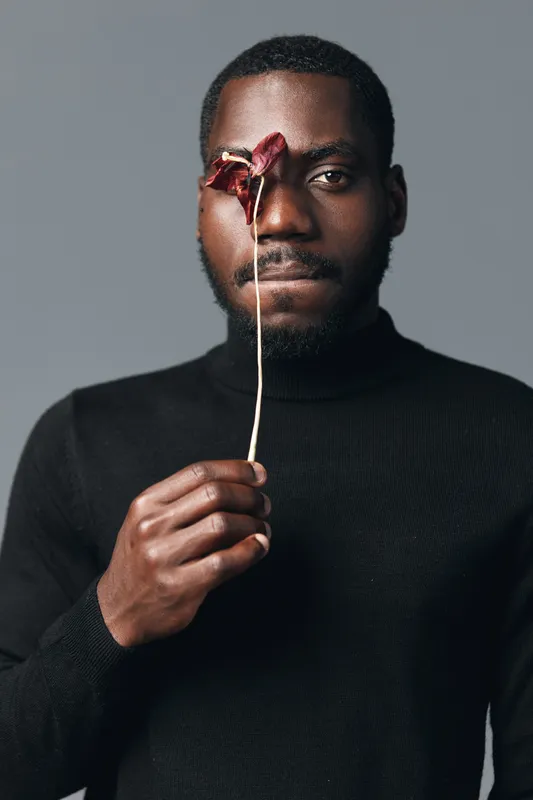 African man with dark skin holding dried flower in front of face in creative conceptual visual style. Portrait of male model wearing black sweater with artistic expression on gray background.