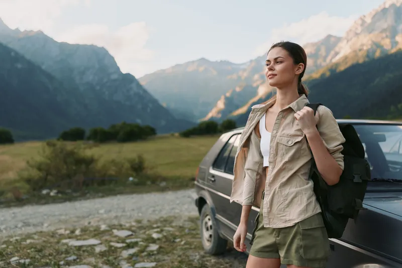 Young woman with a backpack stands beside a car in a mountain valley, ready for a hike with a calm, confident mood. She wears a light shirt and shorts as the landscape opens behind.