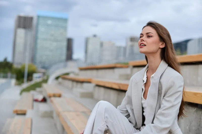 confident woman in light gray suit sitting outdoors in urban business district with modern skyscrapers under cloudy sky