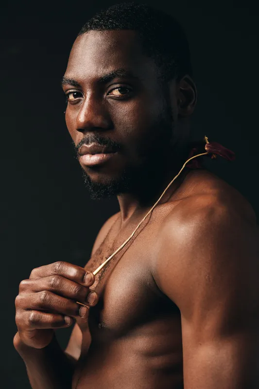 African man with dark skin and muscular body holding a flower in creative conceptual visual style. Portrait of confident male with natural expression and artistic lighting on black background.