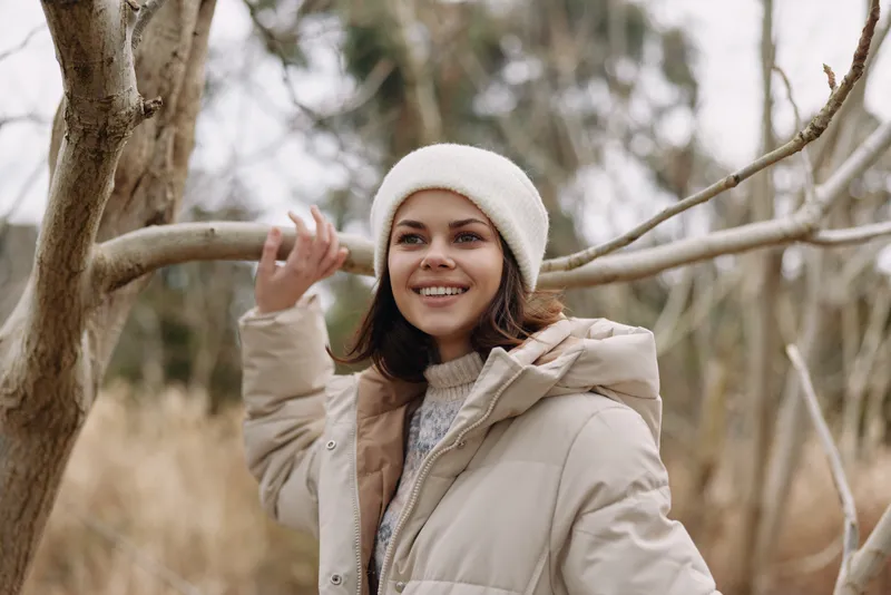 Woman in beige winter jacket and beanie smiles outdoors among trees, capturing cozy outerwear and casual seasonal fashion in a chilly natural setting.