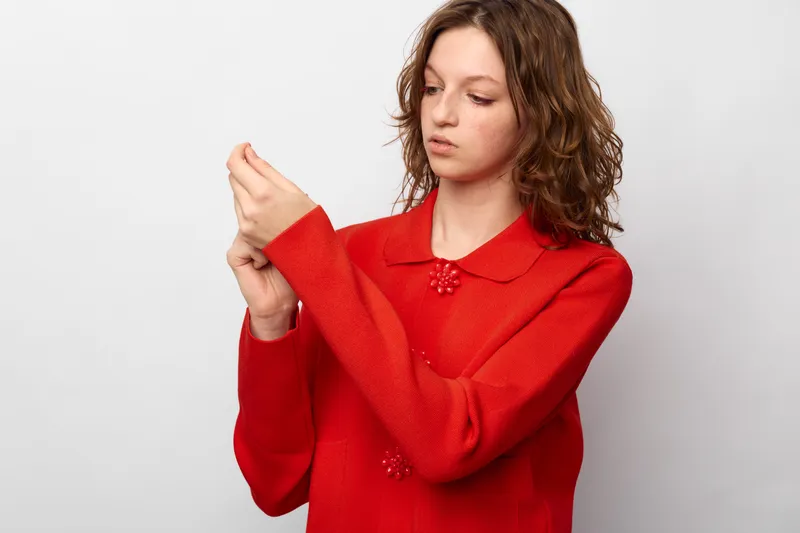 Young woman in red sweater adjusts sleeve with focused expression on white background. Casual fashion, autumn style, and thoughtful mood concept in studio lighting.