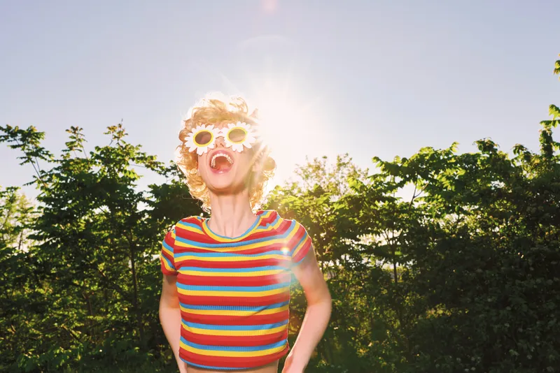 A vibrant woman outdoors in a bright sunny setting wears a striped shirt and flower sunglasses, conveying playful retro fashion and a carefree summer mood, ready for lifestyle campaigns.