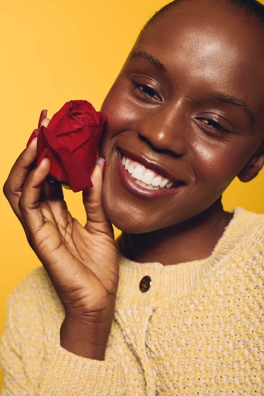 Close-up of smiling black woman holding red rose near face on yellow background. She wears beige sweater, showing joy and natural beauty with dark skin and positive mood concept.