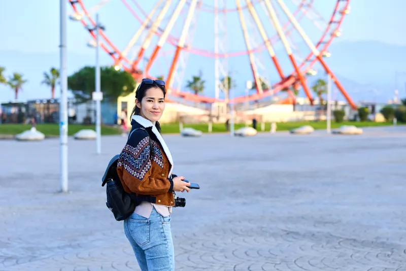 Woman with backpack in a patterned sweater walking in a park near a ferris wheel, outdoors daytime scene, casual style, urban leisure and travel vibe, candid moment.