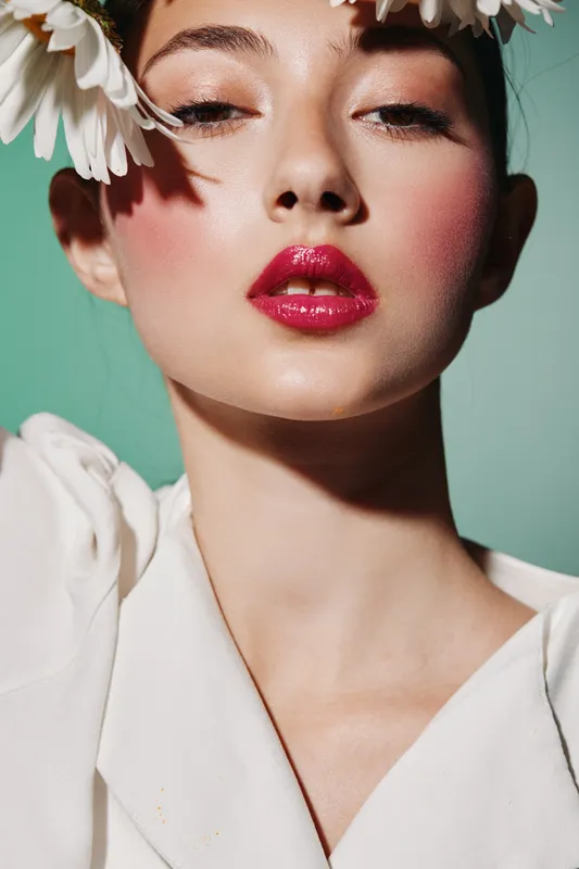 Close-up portrait of a young woman with striking fashion makeup and vivid red lips styled with white flowers on her forehead on green background.