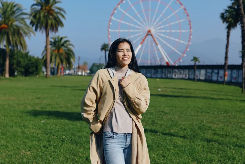 Park scene with a woman in a casual coat enjoying outdoor leisure, standing on green grass with a distant ferris wheel and sunny daylight, radiating a warm smile and relaxed scenery