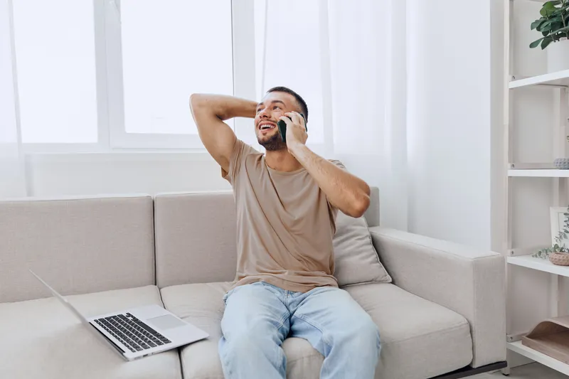 Excited man on a phone call, relaxed at home with a laptop, showcasing modern lifestyle and remote work trends in a bright, airy environment.