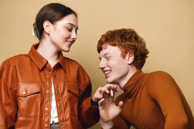 Young man and woman smiling and interacting with warm expressions in casual brown clothing against light neutral background, showing friendship and connection.