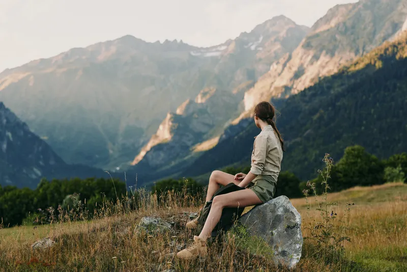 Woman sits on a rock in a mountain meadow, enjoying vast valleys and blue skies. Casual outdoor lifestyle shot emphasizing nature, wellness, and solitude.