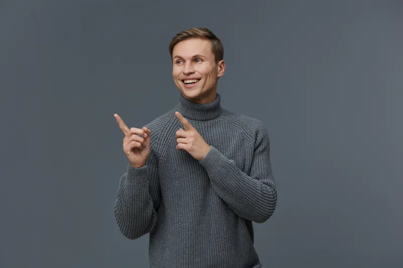 Young smiling happy man wearing grey knitted sweater pointing fingers looking aside isolated on dark background digital detox concept