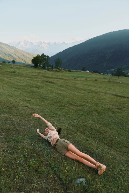 Woman lying on a grassy meadow in a valley with distant mountains, enjoying a carefree outdoor moment, sunlit and relaxed, presenting a natural lifestyle scene in nature.
