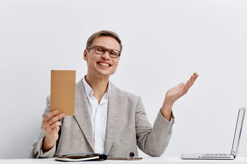 Cheerful smiling man in business attire holding a brown cardboard notebook while sitting at desk with laptop, glasses, and notepad against plain light background. Business, education, presentation con