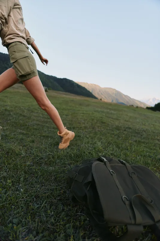 Young woman in khaki shorts leaps across a sunlit meadow with a backpack on the ground, mountains on the horizon, conveying carefree outdoor adventure and movement.