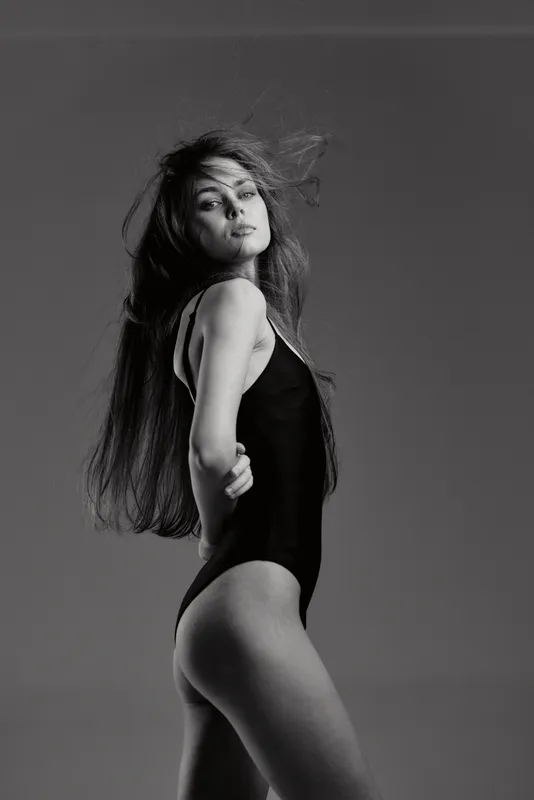 Black and white portrait of a young woman in black bodysuit posing with flowing hair against gray background.