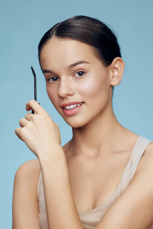 Young woman with brown hair holding a brow tool, smiling against a soft blue background, showcasing natural beauty and wellness in a modern lifestyle concept