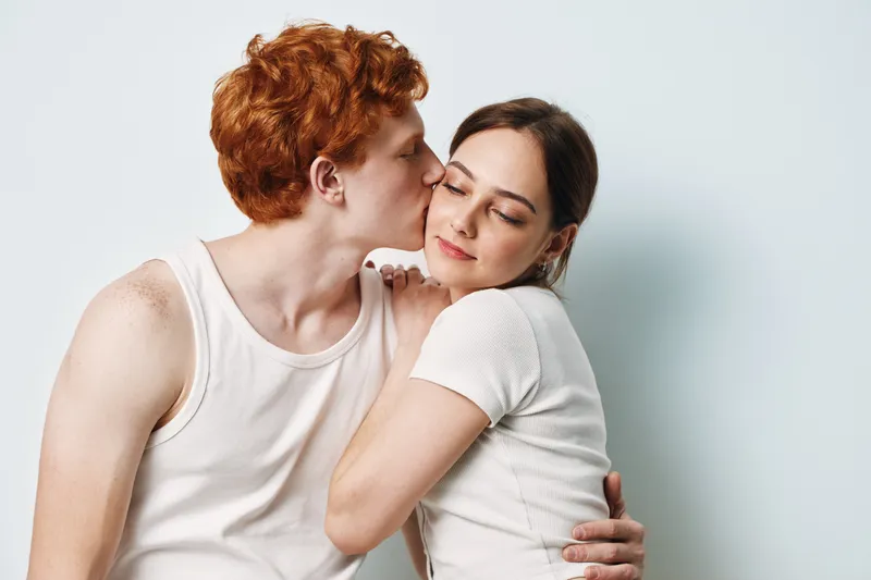 Couple, love, hug and affection with young man kissing woman on cheek in white casual clothes. Relationship, romance and tenderness at indoor studio with light background and skin contact.