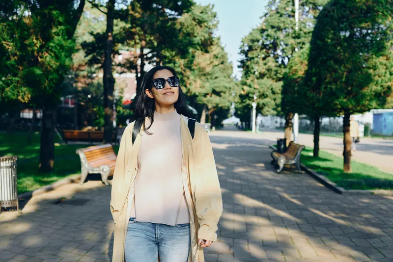 Woman walking along a sunny park path in casual beige cardigan and jeans, wearing sunglasses, enjoying a relaxed outdoor stroll and urban green space.
