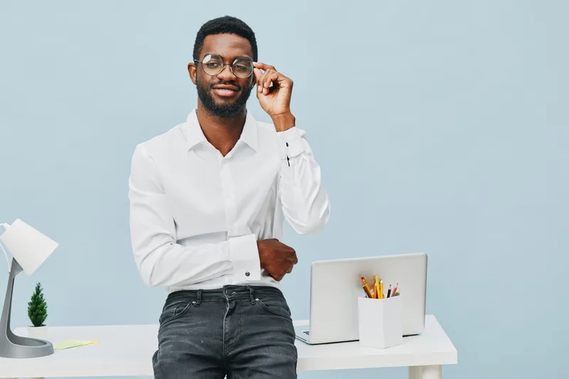 Professional smiling man with glasses in white shirt standing at desk with laptop and stationery on light blue background, studio portrait, business lifestyle concept, confident and approachable appea