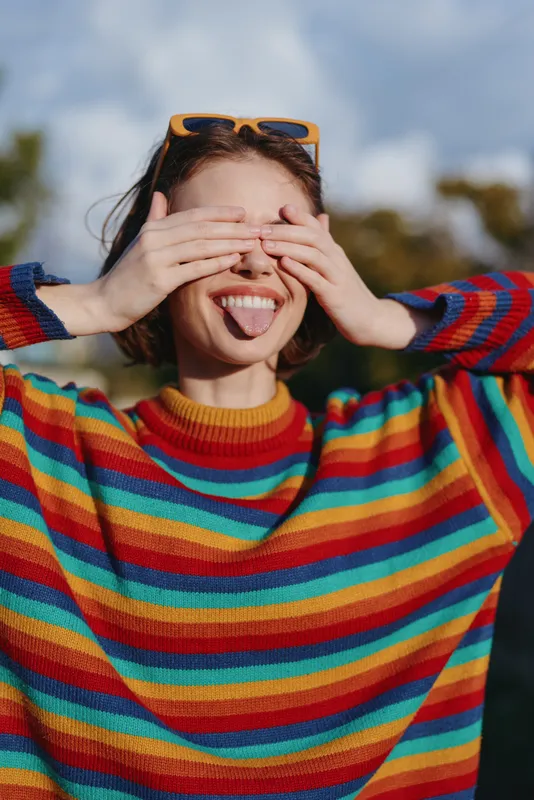 Young woman in her early twenties wearing a rainbow sweater, playful pose in an outdoor portrait, tongue out and sunglasses on head, candid joyful lifestyle moment in park.