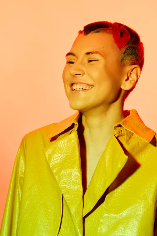 smiling woman with short colorful hair wearing a yellow leather jacket, vibrant orange background, joyful expression, creative fashion portrait, bright lighting