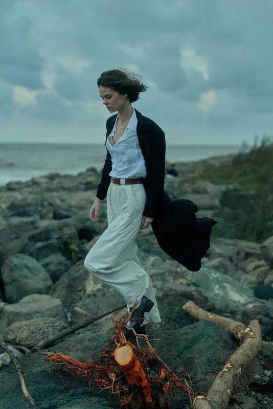 Woman walks along a rocky shoreline by the sea in windy weather, wearing white wide-leg pants, a light blouse, and a dark coat fluttering