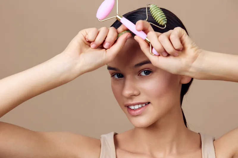 Young woman using facial rollers for skincare, smiling softly with a natural look, pastel background, emphasizing beauty and wellness trends in skincare routines