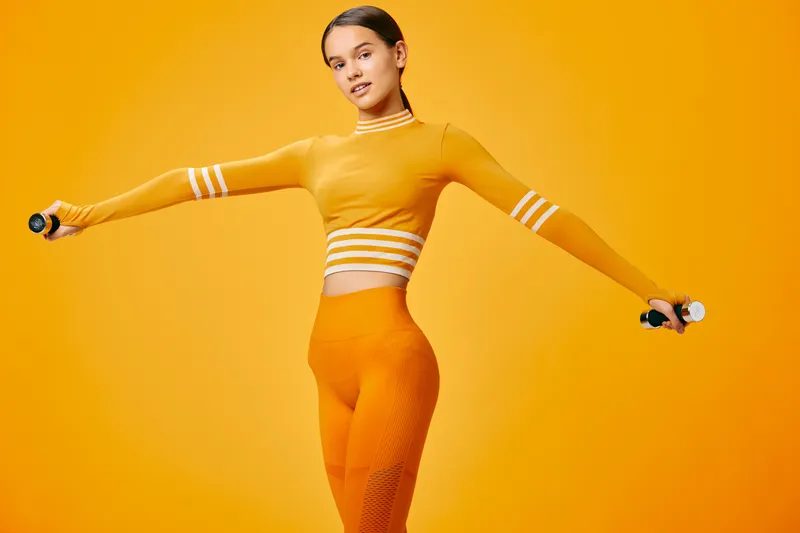 Woman fitness model in bright orange workout outfit holding light dumbbells with outstretched arms, athletic posture and confident expression on a seamless orange studio background.