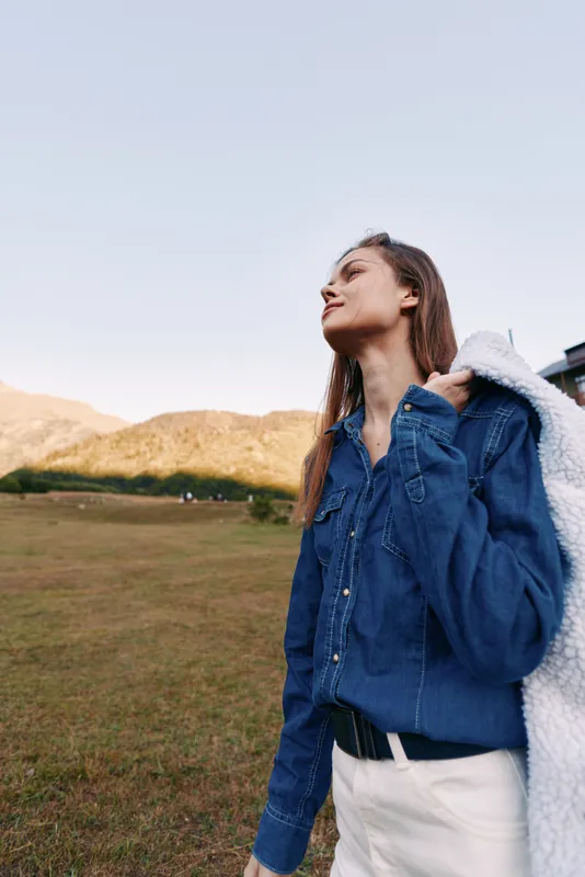 Woman outdoors in a grassy field near mountains, portrait wearing denim shirt and white jacket over shoulder, gazing at sky in a relaxed nature lifestyle scene.