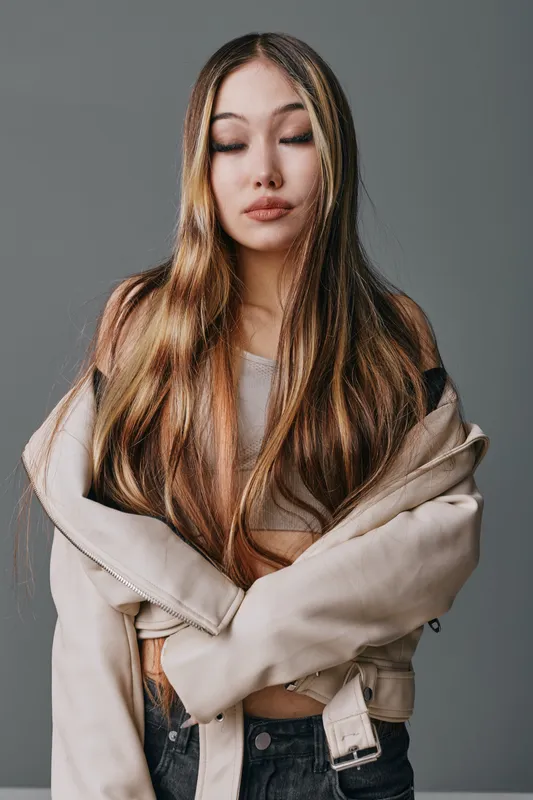 Young woman with long straight hair and closed eyes wearing beige jacket and brown top standing in studio with calm expression and minimalist gray background.