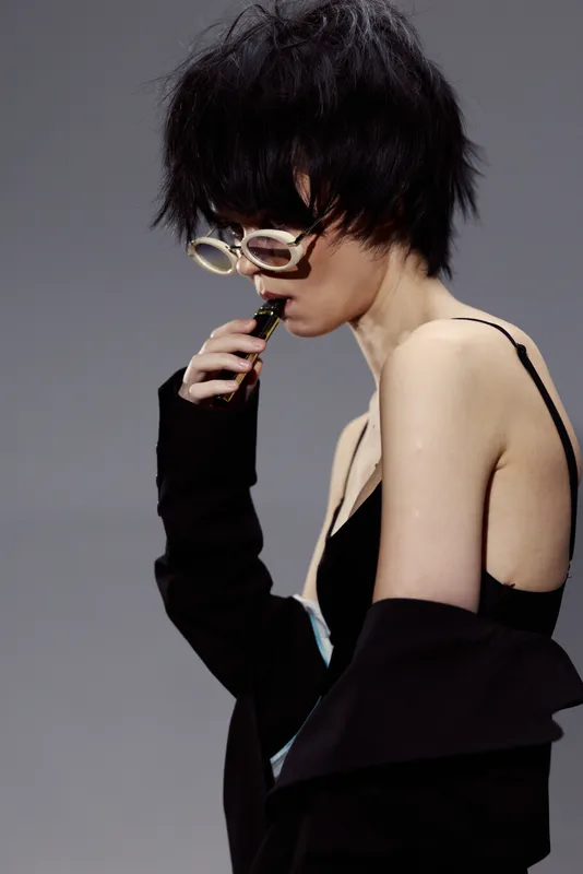 Stylish woman with short black hair wearing white glasses and black clothing in a minimalist gray studio setting