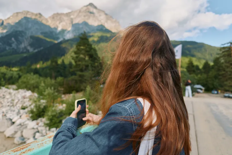 A woman with long brown hair stands outdoors along a scenic mountain road, filming with a smartphone. She wears a denim jacket and smiles, capturing a bright travel moment in nature.