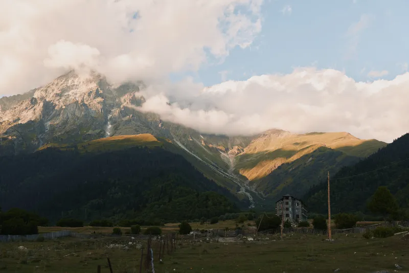 Majestic mountain range under a partly cloudy sky, a quiet rural valley with a lone building and open fields in soft evening light.