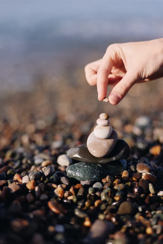 Hands place small smooth stones into a balanced stack on a pebbled beach, capturing focus calm and mindfulness in a natural coastal scene, as waves softly blur in the background