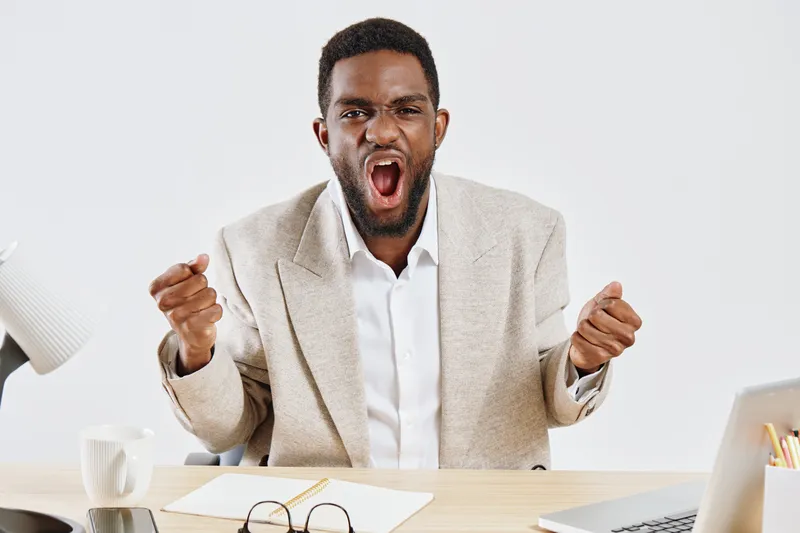 Smiling young man expressing excitement and triumph, wearing beige blazer and white shirt, sitting at desk with laptop and notepad, on plain white background. People lifestyle concept, positive emotio