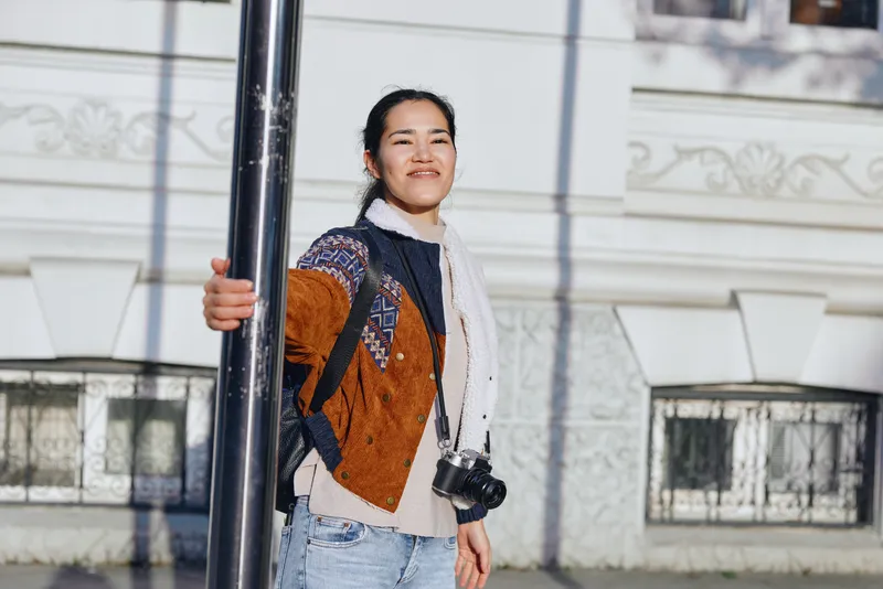 Street scene with a woman posing beside a pole, camera in hand and backpack slung, showcasing urban casual fashion and outdoors lifestyle under bright daylight.