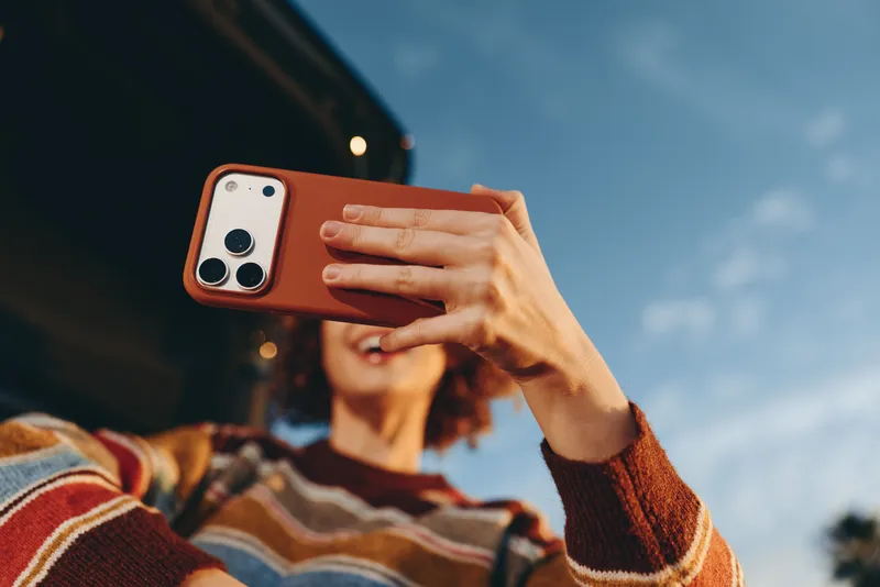 Woman lifestyle outdoors, holding smartphone in her hands, taking photo with orange phone case, casual sweater sleeves visible, sunny sky background, candid selfie moment, today outside