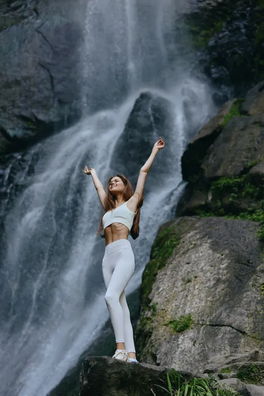 Young woman in white sportswear joyfully stretching near a majestic waterfall surrounded by lush green rocks and natural forest. Energetic outdoor fitness moment.