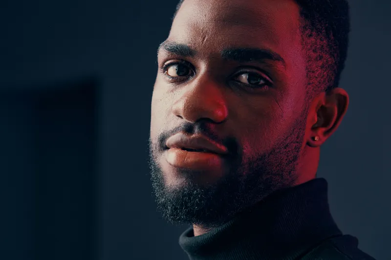 A close up portrait of a young Black man with a stylish beard, wearing a high neck black sweater, expressing confidence in a moody, colorful background