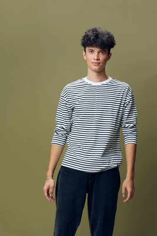 Young man with curly hair wearing a striped long sleeve shirt and dark pants standing against olive green background, casual and relaxed posture.