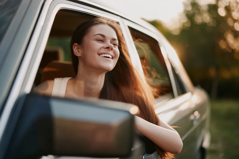 Happy young woman enjoying a drive in a classic car, sun shining, long hair flowing, joyful expression, surrounded by greenery.