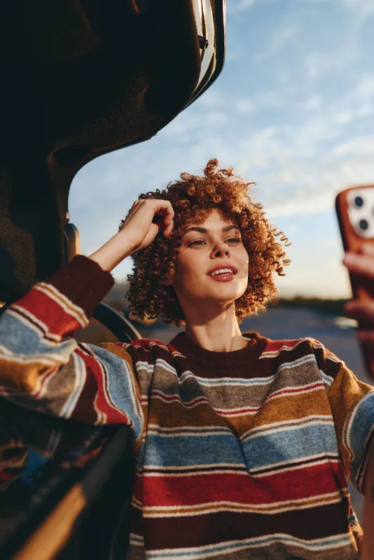 Woman with curly hair adjusting her hair outdoors wearing a striped sweater holding a phone for a selfie in golden light, casual lifestyle, warm happy mood, street background