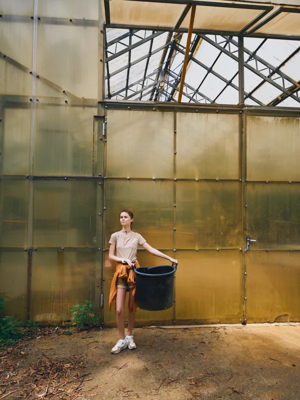 Woman in a greenhouse carrying a black bucket while gardening plants in a casual outfit with sweater and sneakers, standing by translucent glass panels and potted soil in daylight.