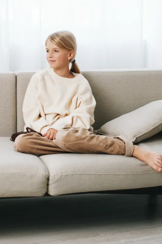 Authentic lifestyle image of a young girl seated on a sofa in a cozy home setting, wearing casual clothes, calm and thoughtful, capturing everyday school life and play moments.