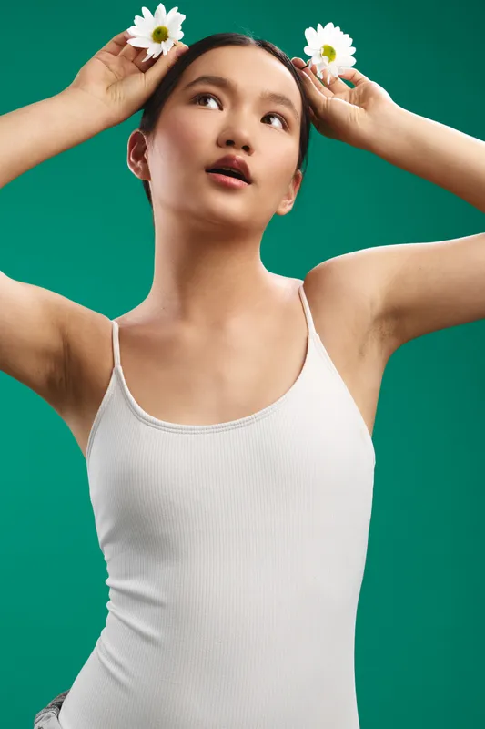 Asian woman poses with small flowers tucked in her hair, standing against a bold green backdrop. She smiles softly, exuding creativity, poise, and a fresh, uplifting vibe in a bright studio setting.